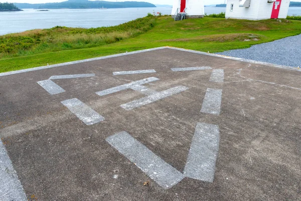 A helicopter landing pad near the ocean. A triangle encloses the letter H, grass, buildings, and water in the background.