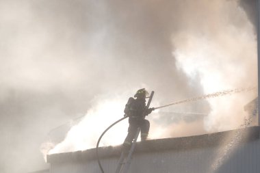 İtfaiyeci merdivenin başında hortumla ateşle savaşıyor. Duman onu kısmen gizliyor. Bir sürü sprey ve duman havayı dolduruyor. Yukarıdaki metne yer.