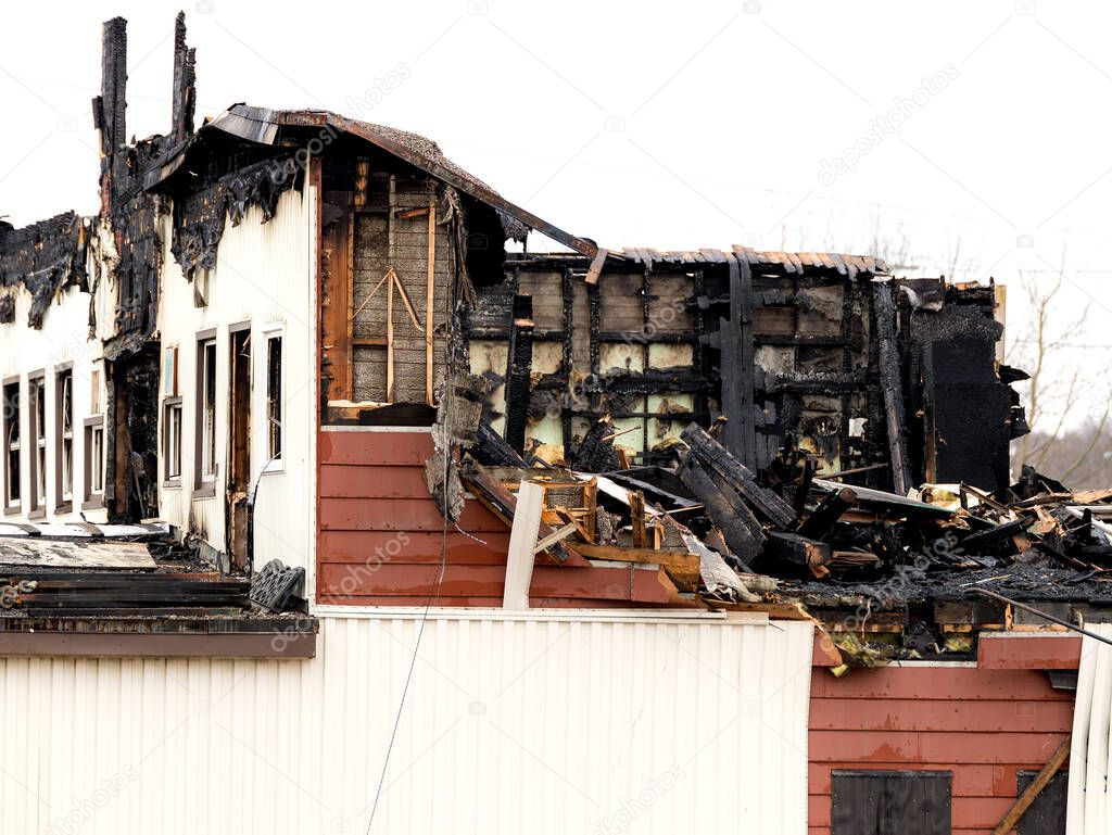 The upper floor of a burned out building. Burned wood and other material is present. The building is partially demolished. Sky is overcast.  Focus is on the closest wall.