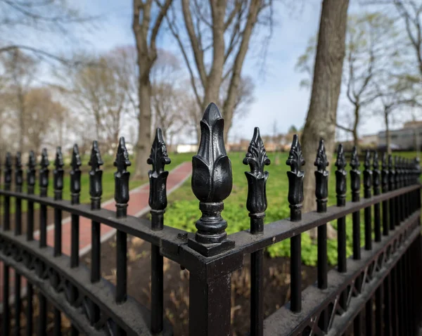 A wrought iron fence by a park. View is towards a corner of the fence. Trees and a brick path are in the background.