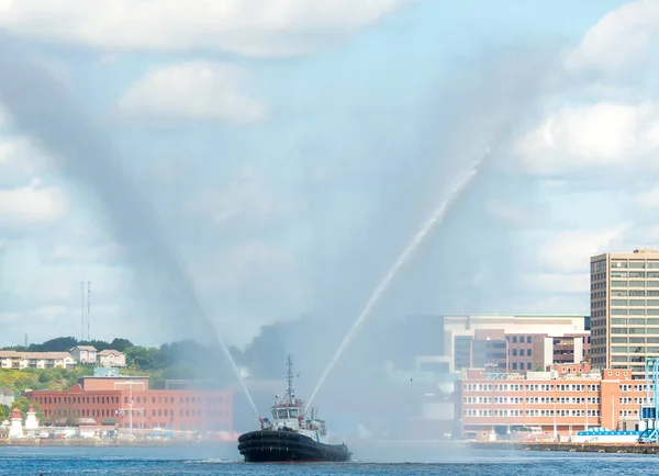Fire tug boat in harbor. Tug is spraying two streams of water into the air in a V shape. Tug partly obscured by spray. City in background. Focus on bow. Identification removed from tug. Room for text.