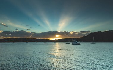 The Esplanade, Ettalong Beach, Central Coast, NSW, Avustralya 'da gündoğumu, botlar ve kreusküler güneş ışınları..