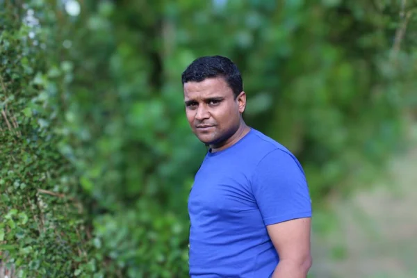 close portrait of young man in blue t shirt standing outside in park with trees in background