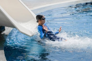Mavi mayo giymiş, mayo takmış, gözlüklü bir çocuk beyaz bir slaytta süzülerek eğleniyor..