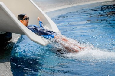 Mavi mayo giymiş, mayo takmış, gözlüklü bir çocuk beyaz bir slaytta süzülerek eğleniyor..