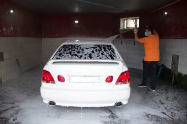 Genç bir adam araba yıkamada suyla beyaz bir araba yıkar.