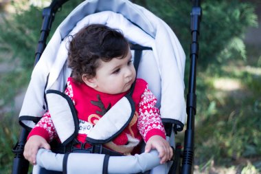 Kırmızı kazaklı küçük çocuk bebek arabasında oturur ve sola bakar.