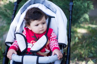 Kırmızı kazaklı küçük çocuk bebek arabasında oturur ve sola bakar.