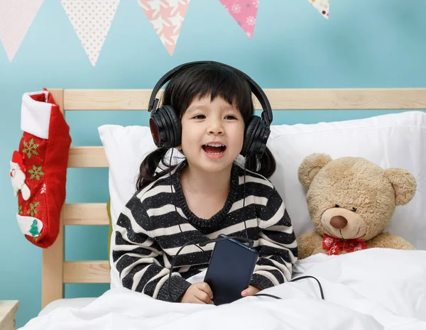 Küçük tatlı bir kız, yatak odasında oyuncak ayıyla birlikte akıllı telefonuyla şarkı söylüyor, Mutlu Asyalı küçük kız kulaklıkla müzik dinliyor, yatağında oyuncak ayı var, teknoloji konsepti var.