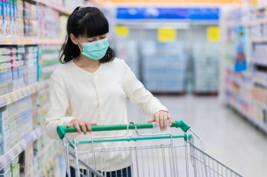 Tıbbi maske takan Asyalı kadın, bakteri ve mikropların yayılmasını önlemek ve korona virüsünden korunmak için süpermarkette alışveriş arabasının sapını siliyor. Hijyen kavramı