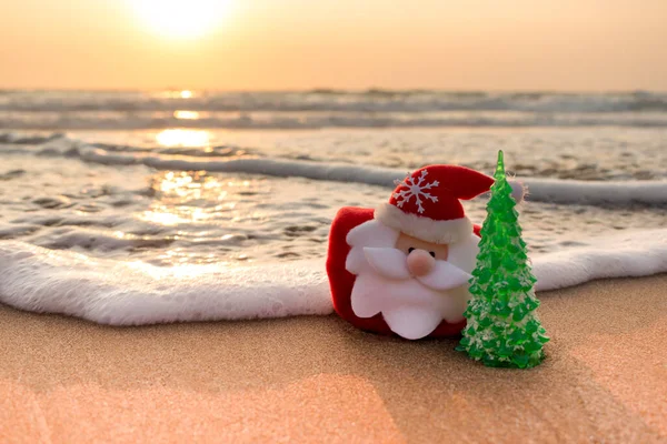 Adriyatik Denizi 'nin kumlu sahilindeki Noel Baba ve Noel ağacı, gün batımının ışığıyla aydınlanan bir dalga arkadan yaklaşıyor. Noel ve Yeni Yıl sıcak ülkelerde seyahat kavramını kutluyor.