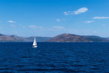 Deniz klasik bir mavi renktir ve üzerinde beyaz bir yelkenli vardır. Yunanistan dağları ve arka planda gökyüzü. İnanılmaz deniz manzarası. Lefkimmi limanından manzara çok güzel. Korfu Adası, Yunanistan.