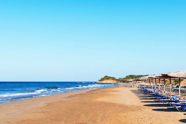 Boş, ıssız bir sahil manzarası. Uzun, kumlu sahil şeridi, mavi deniz, gökyüzü, plaj şemsiyeleri ve boş güverte sandalyeleri. Deniz Burnu. Seyahat, tatil konsepti. Chalikounas Sahili, Korfu Adası. Metin için alanı kopyala