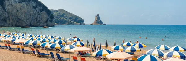 12 Eylül 2019, Agios Gordios plajı, Korfu adası, Yunanistan. Yunan plajının panoramik manzarası - plaj şemsiyeleri ve şezlongları, mavi bir deniz ve gökyüzü ve plajdaki insanlar ile uzun kumlu bir sahil manzarası.