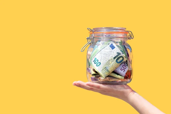 现金欧元钞票在玻璃瓶里 在一个被黄色背景隔离的孩子手里 手握小猪银行的钱 银行和早期规划方面的节约 复制空间 — 图库照片