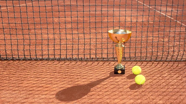 Golden champion trophy and balls on a red clay tennis court and net shadow. Winner in tennis tournament concept. Sports background. Copy space