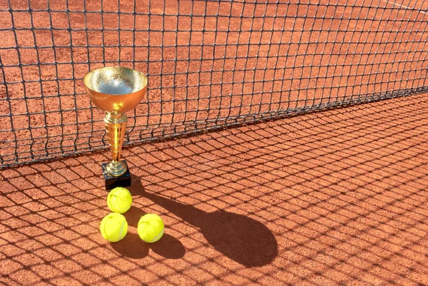 Kupa ve tenis topları kırmızı kil sahada ve ağdan gelen gölgede. Şampiyon ya da zafer kupası. Tenis turnuvasında ya da şampiyonluk konseptinde kazanan. Spor geçmişi. Boşluğu kopyala