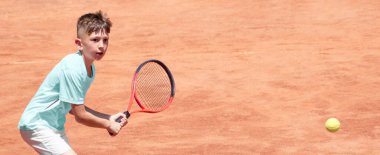 Tenis kortunda elinde raketle çocuk ters vuruş yapmaya hazır. Erkek tenisçi hareket halinde ve hareket halinde. Spor maçı sırasında genç bir sporcunun yüzündeki duygular. Panoramik. Pankart. Boşluğu kopyala
