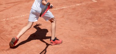 Çocuk kırmızı kil kortunda tenis oynuyor. Tenis kortundaki çocuk forehand vuruyor. Raketi olan genç bir tenisçi. Çocukların gölgeli spor geçmişleri. Sancak boyutu. Boşluğu kopyala