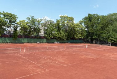 Güneşli yaz gününde boş tenis kortu. Kırmızı kil tenis kortunun, yeşil ağaçların ve arka planda mavi bir gökyüzünün görüntüsü. Tenis için açık hava spor sahası. Metin veya tasarım için boşluk kopyala