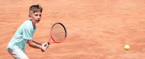 Tenis kortunda elinde raketle çocuk ters vuruş yapmaya hazır. Erkek tenisçi hareket halinde ve hareket halinde. Spor maçı sırasında genç bir sporcunun yüzündeki duygular. Panoramik. Pankart. Boşluğu kopyala