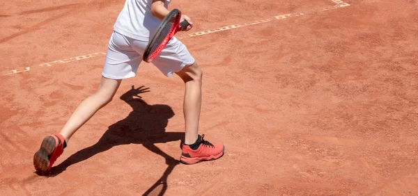 少年は赤い粘土のコートでテニスをする テニスコートの子供は前にヒットします ラケットを持った若いテニス選手が活躍している 子供たちは影と背景をスポーツ バナーサイズ スペースのコピー — ストック写真