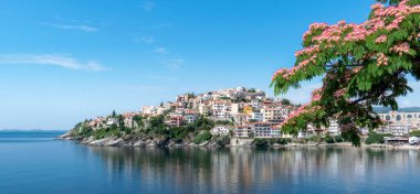 Panoramic view of popular tourist city Kavala, blue sea and sky and blooming Persian silk tree Albizia julibrissin. Beautiful seascape over Aegean Sea, eastern Macedonia, northern Greece. Copy space clipart