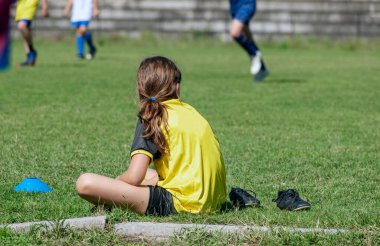 Spor sarı üniformalı tanınmayan bir çocuk futbol sahasının kenarında oturuyor ve arkadaşlarının oynamasını izliyor. Yedek futbolcu takımının maçını izliyor. Seçici Odaklanma. Boşluğu kopyala
