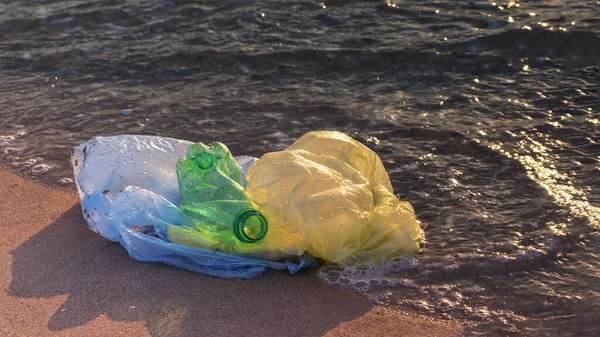Plastic bottle and bags in sea water near shore in golden light of dawn. World ocean pollution. Ecology. Environment protection. Selective focus, copy space