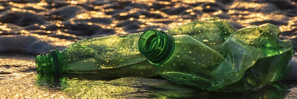 Green plastic bottles in golden glittering ocean waves during sunset. Global environmental problems, plastic pollution of the ocean. Panorama. Banner size