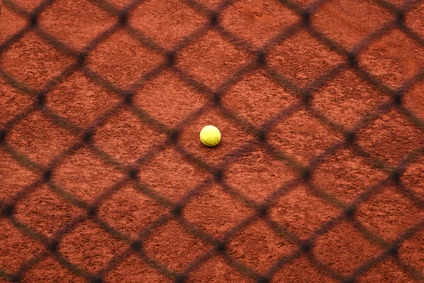 Single tennis ball on red clay tennis court behind the iron fence. Black metal fence and orange clay court behind it - background, texture.