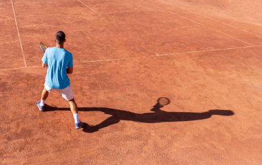 Tenisçi ve gölgesi kırmızı kil tenis kortunda. Profesyonel erkek tenisçi topu almaya hazır. Tenis maçının başlangıcı. Spor geçmişi. Metin için alanı kopyala