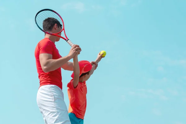 Genç bir tenis koçu ya da eğitmeni bir çocuğa servis tekniğini öğretir. Tenisçi çocuk servis için topu fırlatıyor. Çocuk spor okulu. Mavi gökyüzü arka planı. Pankart. Metin için alanı kopyala.