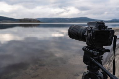 Arka planda bulanık Batak gölü ve dağ manzarası olan tripodun üzerindeki kameranın yakın çekimi, fotoğraf ekipmanlarına odaklan