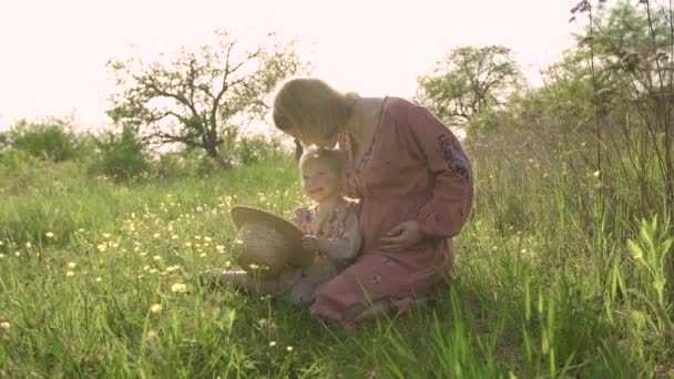 Mała dziewczynka w sukience trzyma słomkowy kapelusz. Ciężarna matka w haftowanej sukience całuje swoją 2-letnią córkę w głowę. Mama i córka siedzą na trawie w ogrodzie i bawią się — Wideo stockowe