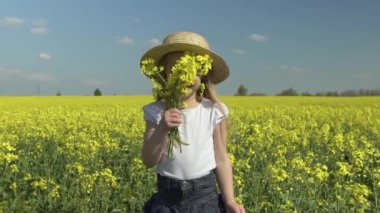 Hasır şapkalı küçük güzel mutlu kız sarı bir tarlada gülümser ve bir buket sarı çiçeğin arkasına saklanır, ağır çekimde.