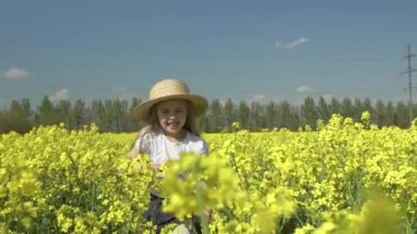 Sarı saçlı, hasır şapkalı küçük mutlu kız sarı bir tarlada koşar ve gülümser, ağır çekimde.