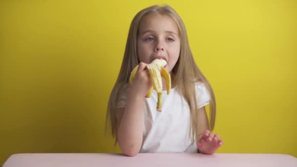 노란 배경에 흰색 티셔츠를 입은 금발의 아름다운 소녀가 바나나를 먹고 그것을 바라봅니다., — 비디오