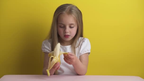 흰색 티셔츠를 입은 금발의 아름다운 소녀가 노란 배경에 바나나 껍질을 두르고 있다. 바나나를 먹고 미소짓습니다. 행복 한 어린 시절, — 비디오