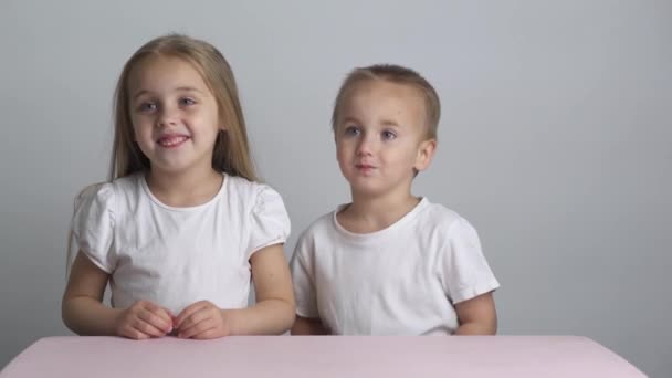 白いTシャツと金髪の髪の白人の妹と弟がテーブルに座っている。お姉ちゃん抱擁兄とキス — ストック動画