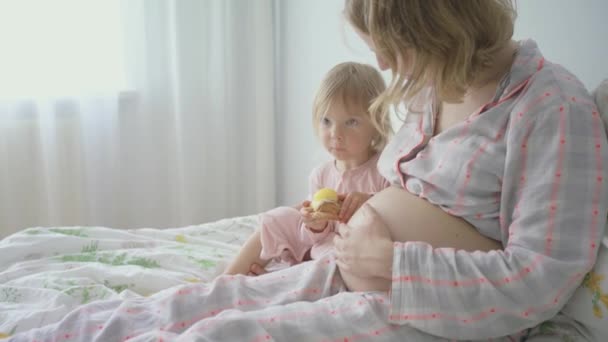 Nahaufnahme. Schwangere Frau im Schlafanzug mit ihrer Tochter im Bett, die ein Croissant mit Öko-Utensilien isst — Stockvideo