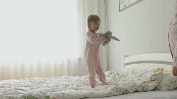 잠옷입은 백인 여자 애가 침대에서 뛰어내렸어요. 장난감 토끼와 재미있는 게임을 하는 모습. — 비디오