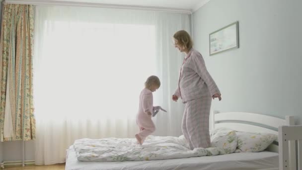 妊娠中の母親とパジャマ姿の娘が寝室のベッドでジャンプします。家での幸せな時間. — ストック動画