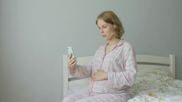 Uma mulher grávida de pijama na cama olha para o telefone e pratica técnicas de respiração — Vídeo de Stock