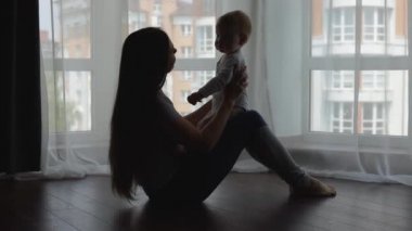 Anne ve oğlu büyük bir pencerenin arka planında, ahşap bir zeminde oturuyor..