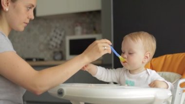 Orta ölçekli. Annem küçük bir çocuğu kaşıkla besler. Küçük bir çocuk beslenme koltuğunda oturur ve yemek yer.