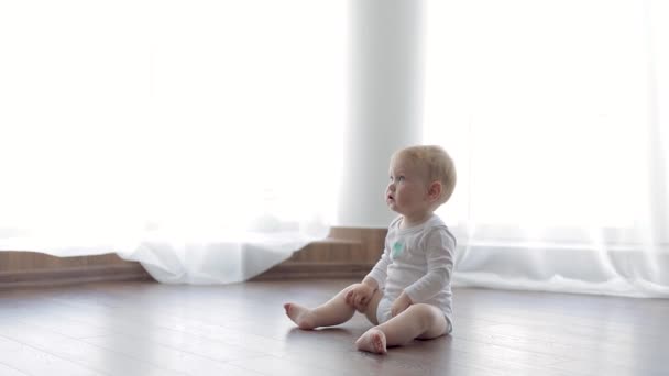 Little baby in a white bodysuit sitting on a wooden warm floor. — Stock Video