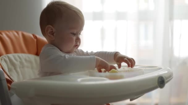 サイドビュー。接近中だ。小さな男の子は餌やりの椅子で彼の手で赤ちゃんの食べ物を食べる — ストック動画