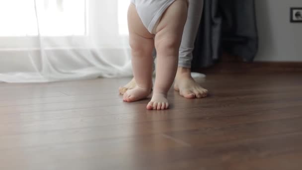 De eerste stappen. Een klein kind leert lopen terwijl het zijn moeders handen vasthoudt. Kaukasisch kind zet zijn eerste stappen op de houten verwarmde warme vloer — Stockvideo
