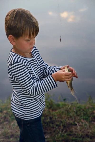 少年は魚を持っており 日没時に村の湖を背景に立っています — ストック写真